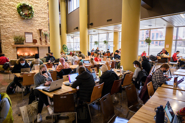 A welcoming fireplace and holiday wreath are seen in the background as students on laptops study at the Union South atrium.